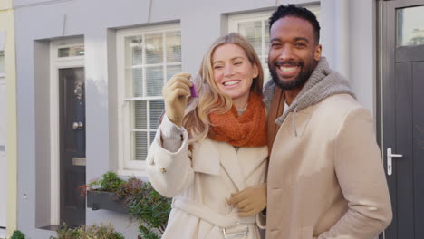 Portrait-of-multi-cultural-couple-outside-house-on-moving-day-holding-keys-to-new-home-in-fall-or-winter---shot-in-slow-motion
