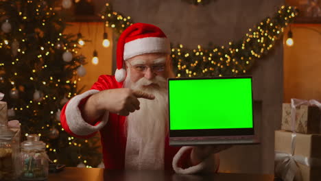 Im-Hintergrund-Des-Weihnachtsbaums-Hält-Ein-älterer-Weihnachtsmann-Einen-Laptop-Und-Zeigt-Mit-Einem-Chromakey-Auf-Den-Bildschirm.-Laptop-Mit-Grünem-Bildschirm.-Der-Weihnachtsmann-Schaut-In-Die-Kamera.-Hochwertiges-4K-Filmmaterial