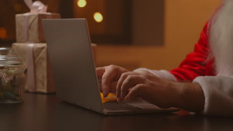 Hände-Des-Weihnachtsmanns-Tippen-Auf-Einer-Kabellosen-Tastatur-Neben-Einem-Mit-Neujahr-Dekorierten-Holztisch.-Der-Weihnachtsmann-Arbeitet-Mit-Einem-Laptop,-Sieht-E-Mails-Durch-Und-Beantwortet-Nachrichten-An-Kinder.-Hochwertiges-4K-Filmmaterial