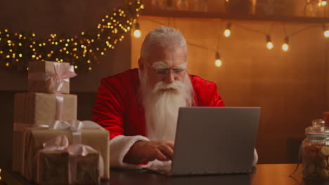 Papá-Noel-Real-Que-Utiliza-Nueva-Tecnología-Para-Comunicarse-Con-Los-Niños-Que-Reciben-Correo-O-Lista-De-Deseos.-Alegre-Trabajando-En-Una-Computadora-Portátil-Y-Sonriendo-Mientras-Está-Sentado-En-Su-Silla-Con-Chimenea-Y-árbol-De-Navidad.-Imágenes-4k-De-Alta-Calidad