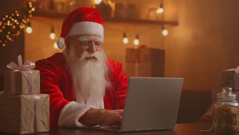 Papá-Noel-Sosteniendo-Una-Caja-De-Regalo,-Escribiendo-Un-Teclado,-Un-Niño-Hablando-Con-Un-Niño-Saludando-En-Feliz-Navidad,-Feliz-Año-Nuevo,-En-Un-Chat-Virtual-En-Línea-En-Una-Computadora-Portátil-Sentado-En-La-Mesa-De-Casa-Tarde-Con-Un-Regalo-En-La-Víspera-De-Navidad.-Imágenes-4k-De-Alta-Calidad