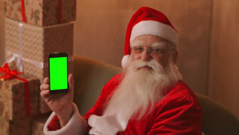 Santa-Claus-is-sitting-on-the-sofa-in-the-background-of-a-Christmas-tree-and-garlands-holding-a-mobile-phone-with-a-green-screen-pointing-at-it-with-his-finger.-Santa-holds-a-phone-with-a-chromakey-on-the-screen