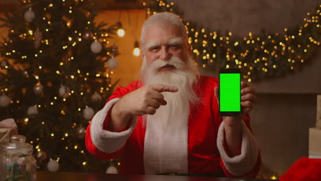 Papá-Noel-Está-Sentado-En-El-Sofá-Al-Fondo-De-Un-árbol-De-Navidad-Y-Guirnaldas-Sosteniendo-Un-Teléfono-Móvil-Con-Una-Pantalla-Verde-Apuntándolo-Con-El-Dedo.-Santa-Sostiene-Un-Teléfono-Con-Un-Cromakey-En-La-Pantalla