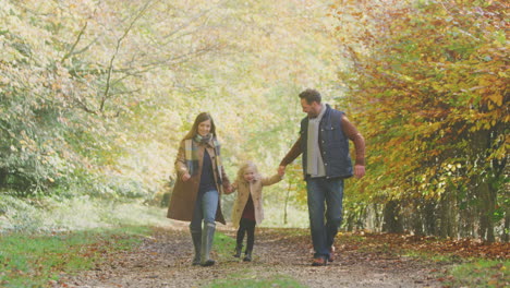 Excited-Girl-Being-Swung-By-Parents-On-Family-Autumn-Walk-Through-Countryside