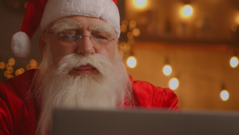 Weihnachtsmann-Benutzt-Laptop-Im-Wohnzimmer.-Porträt-Des-Nachdenklichen-Weihnachtsmanns,-Der-Mit-Modernem-Laptop-In-Dekoriertem-Haus-Tippt.-Hochwertiges-4K-Filmmaterial