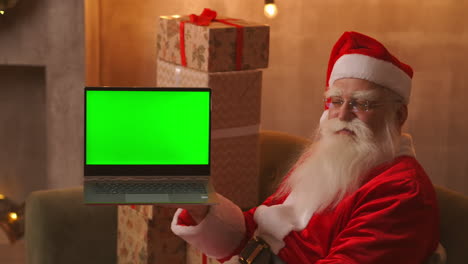 En-El-Fondo-Del-árbol-De-Navidad,-Un-Anciano-Papá-Noel-Sostiene-Una-Computadora-Portátil-Y-Apunta-A-La-Pantalla-Con-Una-Croma.-Portátil-Con-Pantalla-Verde.-Papá-Noel-Mira-A-La-Cámara.-Imágenes-4k-De-Alta-Calidad