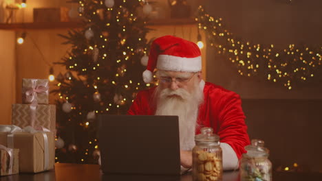 Santa-Claus-holding-gift-box-typing-keyboard-kid-talking-to-child-greeting-on-Merry-Christmas-Happy-New-Year-in-virtual-online-chat-on-laptop-sitting-at-home-table-late-with-present-on-xmas-eve.-High-quality-4k-footage