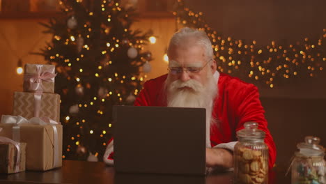 Santa-Claus-holding-gift-box-typing-keyboard-kid-talking-to-child-greeting-on-Merry-Christmas-Happy-New-Year-in-virtual-online-chat-on-laptop-sitting-at-home-table-late-with-present-on-xmas-eve.-High-quality-4k-footage