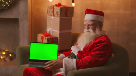 In-the-background-of-the-Christmas-tree-an-elderly-Santa-Claus-is-holding-a-laptop-and-pointing-at-the-screen-with-a-chromakey.-Laptop-with-a-green-screen.-Santa-looks-at-the-camera.-High-quality-4k-footage