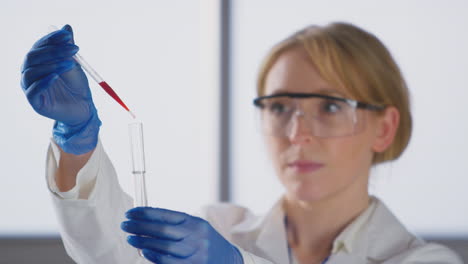 Female-Lab-Worker-Wearing-White-Coat-Adding-Red-Liquid-From-Pipette-Into-Test-Tube