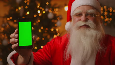 Papá-Noel-Está-Sentado-En-El-Sofá-Al-Fondo-De-Un-árbol-De-Navidad-Y-Guirnaldas-Sosteniendo-Un-Teléfono-Móvil-Con-Una-Pantalla-Verde-Apuntándolo-Con-El-Dedo.-Santa-Sostiene-Un-Teléfono-Con-Un-Cromakey-En-La-Pantalla