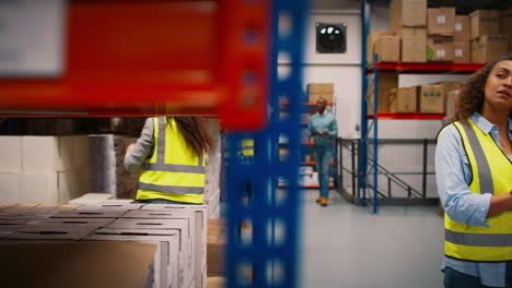 Team-Leader-With-Digital-Tablet-In-Busy-Warehouse-Training-Female-Intern-Standing-By-Shelves