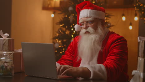 Papá-Noel-Sosteniendo-Una-Caja-De-Regalo,-Escribiendo-Un-Teclado,-Un-Niño-Hablando-Con-Un-Niño-Saludando-En-Feliz-Navidad,-Feliz-Año-Nuevo,-En-Un-Chat-Virtual-En-Línea-En-Una-Computadora-Portátil-Sentado-En-La-Mesa-De-Casa-Tarde-Con-Un-Regalo-En-La-Víspera-De-Navidad.-Imágenes-4k-De-Alta-Calidad