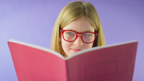 Studioaufnahme-Eines-Jungen-Mädchens-Mit-Brille,-Das-Schulhefte-Vor-Violettem-Hintergrund-Studiert