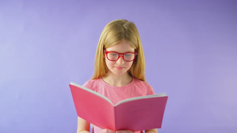 Studioaufnahme-Eines-Jungen-Mädchens-Mit-Brille,-Das-Schulhefte-Vor-Violettem-Hintergrund-Studiert