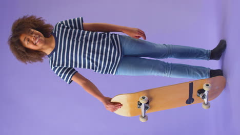 Vertikales-Video-Eines-Kleinen-Jungen,-Der-Vor-Violettem-Hintergrund-Steht-Und-Ein-Skateboard-Hält
