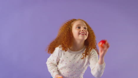 Studioporträt-Eines-Mädchens,-Das-Apfel-Und-Orange-Vor-Violettem-Hintergrund-Jongliert