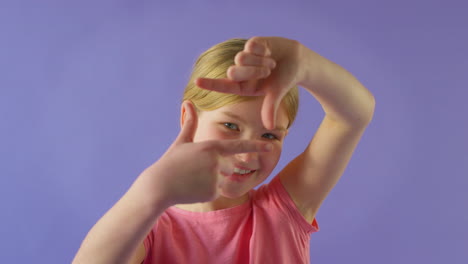 Studio-Portrait-Of-Smiling-Girl-Making-Shape-Of-Picture-Frame-With-Hands-Against-Purple-Background