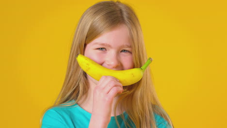 Studioporträt-Eines-Mädchens,-Das-Eine-Banane-Für-Einen-Lächelnden-Mund-Vor-Gelbem-Hintergrund-Hält