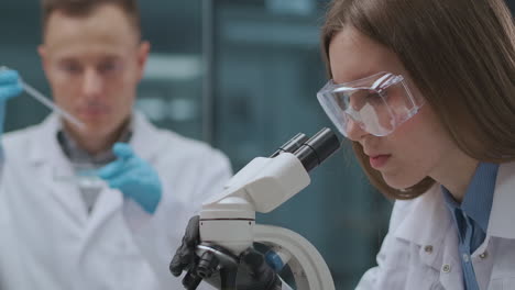 Los-Microbiólogos-Están-Investigando-Virus-Y-Bacterias-En-El-Laboratorio-La-Mujer-Está-Viendo-Por-Microscopio-El-Hombre-Está-Probando-Medicamentos-En-Tubos-De-Ensayo-En-El-Centro-Médico