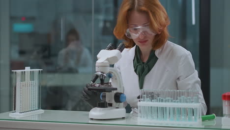 Una-Mujer-De-Mediana-Edad-Está-Trabajando-En-Un-Laboratorio-Criminalístico-Investigando-Pruebas-Con-Un-Microscopio.-Los-Tubos-De-Ensayo-Están-Parados-En-La-Mesa-De-Análisis-De-ADN-En-El-Laboratorio.