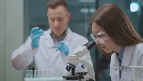 Un-Hombre-Y-Una-Mujer-Están-Trabajando-En-Un-Laboratorio-Médico,-Un-Especialista-Masculino-Está-Probando-Productos-Químicos-En-Tubos-De-Ensayo-Y-Una-Experta-Está-Viendo-Muestras-En-El-Microscopio