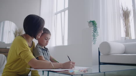 Mother-in-yellow-jacket-and-son-in-t-shirt-sit-at-the-table-and-draw-together-color-pencils-on-paper.-Happy-childhood.-Loving-helps-his-son-in-pre-school-training-and-develops-creativity-in-the-child