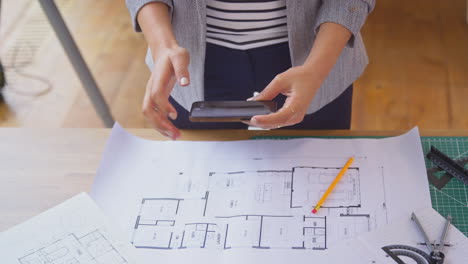 Female-Architect-Working-In-Office-Sitting-At-Desk-Taking-Picture-Of-Plans-On-Mobile-Phone