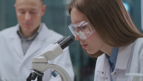 El-Equipo-De-Expertos-Forenses-Está-Trabajando-En-Un-Laboratorio-Criminalístico-Investigando-Evidencia.-La-Mujer-Está-Viendo-Muestras-De-ADN-Con-Un-Microscopio.-El-Químico-Masculino-Está-Explorando-Reactivos.