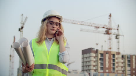 Woman-engineer-designer-talking-on-the-phone-with-the-contractor-with-drawings-in-hand-on-the-background-of-buildings-under-construction-and-cranes