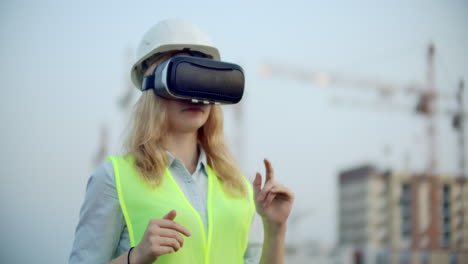 Woman-designer-on-a-building-site-in-hard-hat-and-vest-in-the-glasses-of-virtual-reality-to-move-your-hands-mimicking-the-interface-on-the-background-of-the-cranes-at-sunset.