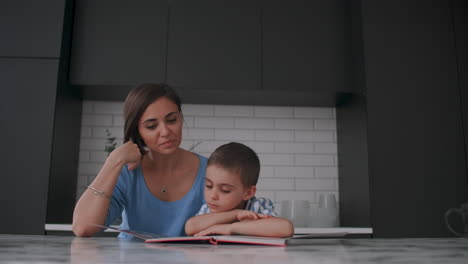 Joven-Madre-Española-E-Hijo-Sentados-En-La-Mesa-Leyendo-Un-Libro-Juntos-Y-Sonriendo