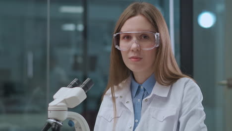 young-woman-technician-lab-is-working-with-microscope-and-looking-to-camera-portrait-of-female-health-professional-in-hospital-or-science-Institute