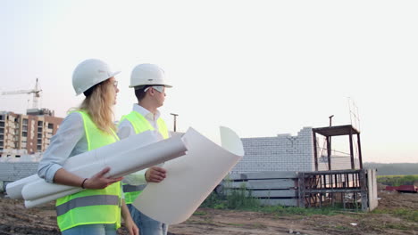 Der-Bauunternehmer-Und-Der-Architekt-–-Mann-Und-Frau-–-Besprechen-Den-Bauplan-Des-Modernen-Geschäftszentrums,-Das-In-Der-Nähe-Der-Baustelle-Steht.-Sie-Gehen-Zum-Gebäude.