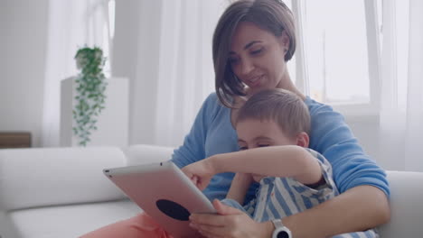 Madre-E-Hijo-Usando-Una-Tableta-Mirando-La-Pantalla-Y-Hablando-En-Un-Sofá-En-Una-Casa-Acogedora