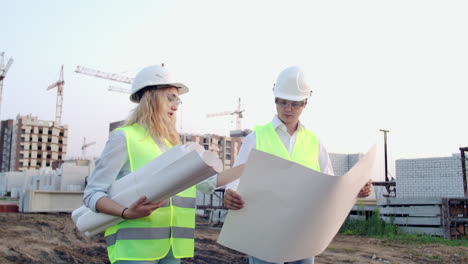 Trabajadores-Con-Dibujos-En-El-Sitio-De-Construcción.-Dos-Trabajadores,-Hombre-Y-Mujer,-Con-Sombreros-Protectores-Trabajando-Con-Dibujos-En-El-Sitio-De-Construcción-Al-Aire-Libre.