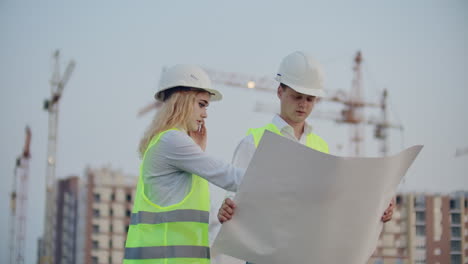 Dos-Ingenieros-Discuten-Observando-Los-Dibujos-Del-Plan-De-Construcción-Y-La-Ubicación-De-Los-Objetos,-Especificando-Los-Detalles-Del-Contratista-En-El-Teléfono.-Hablando-Por-Teléfono-En-El-Sitio-De-Construcción.