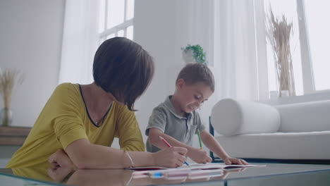 Mother-in-yellow-jacket-and-son-in-t-shirt-sit-at-the-table-and-draw-together-color-pencils-on-paper.-Happy-childhood.-Loving-helps-his-son-in-pre-school-training-and-develops-creativity-in-the-child