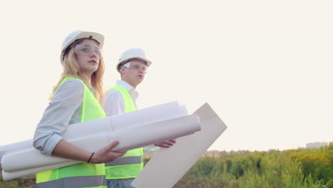 Ein-Bauarbeiter-Und-Eine-Bauarbeiterin-Mit-Zeichnungen-In-Weißen-Helmen-Gehen-Auf-Die-Baustelle-Und-Unterhalten-Sich