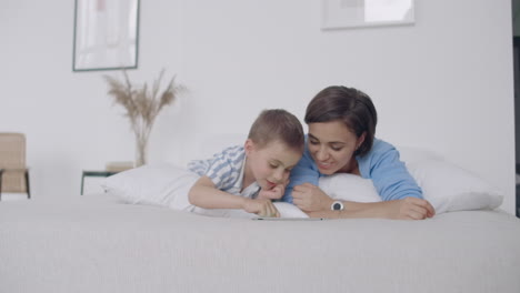 Mom-and-son-looking-at-the-tablet-screen-lying-on-a-white-bed.-Play-games-with-your-son-on-your-tablet-computer-and-watch-funny-videos.