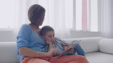 Glückliche-Familie,-Mutter-Und-Kleiner-Sohn-Benutzen-Ein-Digitales-Tablet,-Sitzen-Auf-Dem-Sofa,-Lächelnde-Eltern,-Mutter-Und-Kleiner-Sohn-Halten-Einen-PC-Und-Schauen-Auf-Den-Bildschirm,-Machen-Online-Shopping,-Tätigen-Videoanrufe,-Sehen-Sich-Zeichentrickfilme-An