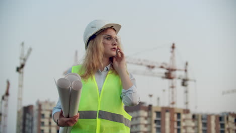 Retrato-De-Una-Ingeniera-Con-Casco-Hablando-Por-Teléfono-En-El-Fondo-De-La-Construcción-Con-Grúas-Sosteniendo-Dibujos-En-Sus-Manos.-Ingeniera-En-El-Sitio-De-Construcción.