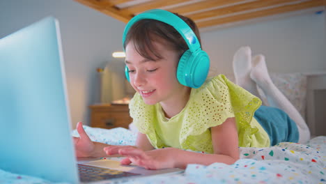 Girl-In-Bedroom-Lying-On-Bed-Wearing-Wireless-Headphones-Using-Laptop-Computer