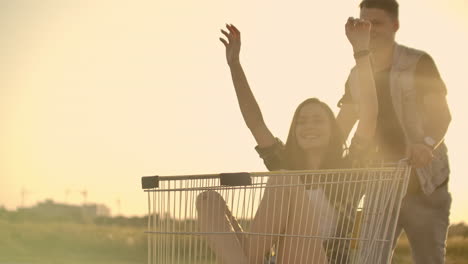 Side-view-of-a-young-man-and-woman-having-fun-outdoors-on-shopping-trolleys.-Multiethnic-young-people-racing-on-shopping-carts.-On-the-parking-zone-with-their