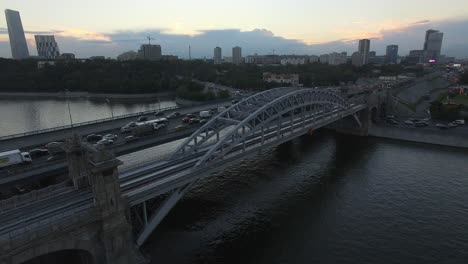 Una-Vista-Aérea-De-Un-Puente-Ferroviario-Sobre-El-Río-Contra-La-Vista-Urbana-Nocturna