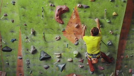 Male-climber-climbs-the-cliff-wall-in-the-room-reaching-and-gripping-hold.-A-professional-concentrated-climber-moves-up-the-wall-the-camera-rises-and-lays-down-his-movements.-Boom-and-truck-follow-camera.