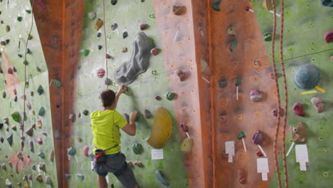 Male-climber-climbs-the-cliff-wall-in-the-room-reaching-and-gripping-hold.-A-professional-concentrated-climber-moves-up-the-wall-the-camera-rises-and-lays-down-his-movements.-Boom-and-truck-follow-camera.