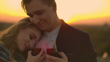 Man-and-young-pretty-woman-sitting-on-couch-holding-wine-glasses-and-kissing-on-rooftop-terrace-at-sunset