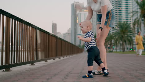 Happy-cute-baby-learns-to-walk-and-takes-first-steps-in-the-city-under-the-control-of-his-mom-holding-baby's-hands-in-the-background-of-the-city