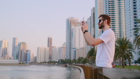 Ein-Junger-Mann-In-Einem-Weißen-T-Shirt-Steht-Am-Wasser-Vor-Dem-Hintergrund-Der-Modernen-Stadt-Und-Macht-Fotos-Und-Videos-Mit-Dem-Telefon